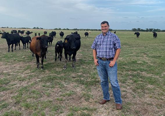 Scott Reed dressed in blue jeans and a blue plaid shirt stands in a pasture with cows in the background.