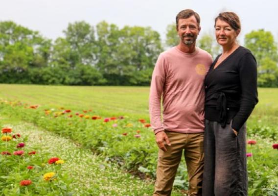 A man and woman stand next to each other on a field with rows of flowers planted behind them.