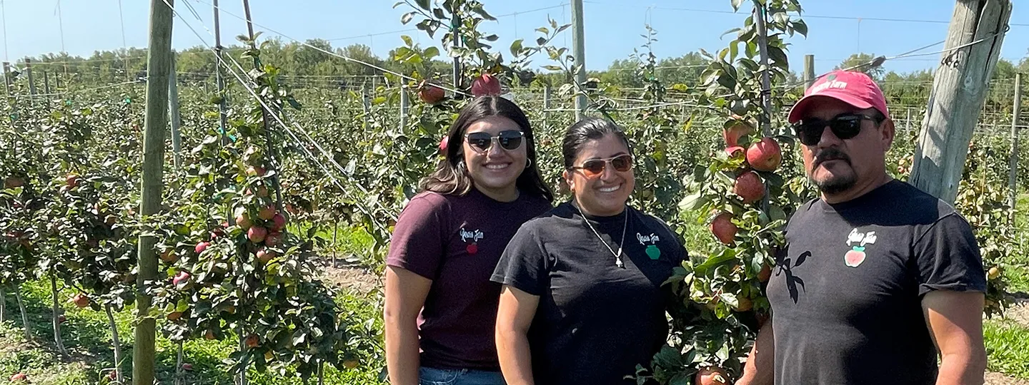 Three farmers stand in front of apples in their orchard